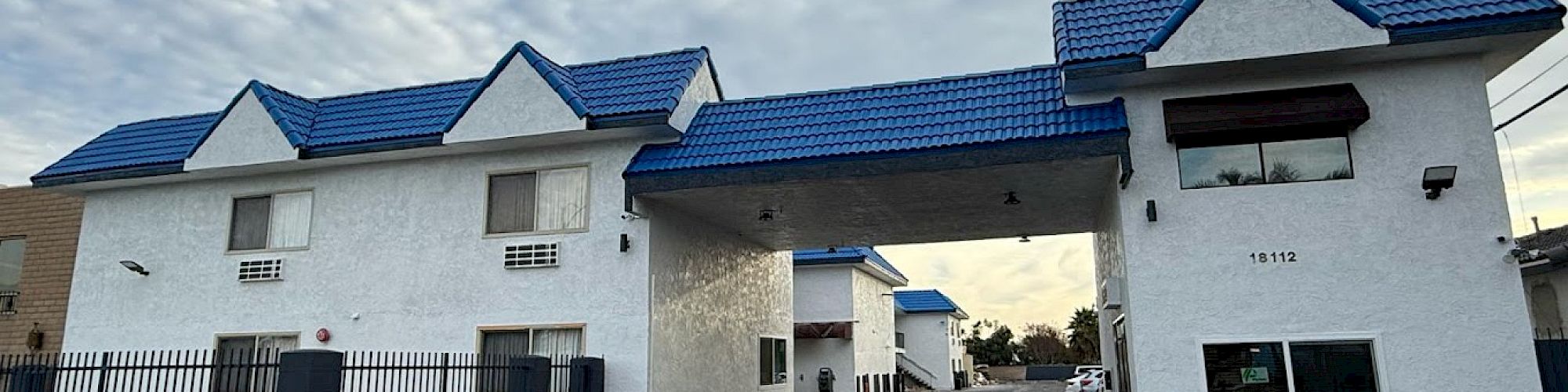 The image shows a white building with blue roofs, featuring an archway entrance. There's a fenced area and parking spaces visible in front.