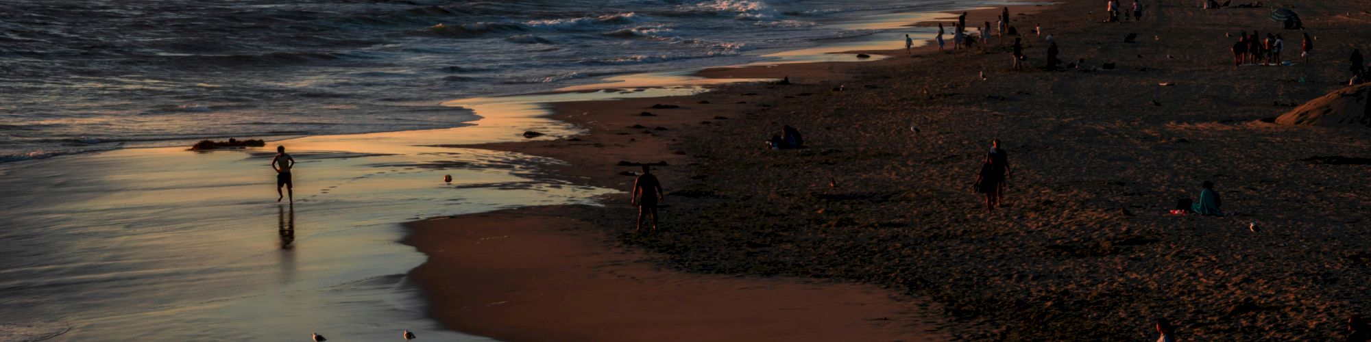 A serene beach scene at sunset with people strolling along the shoreline, water reflecting the sky, and palm trees in the distance.