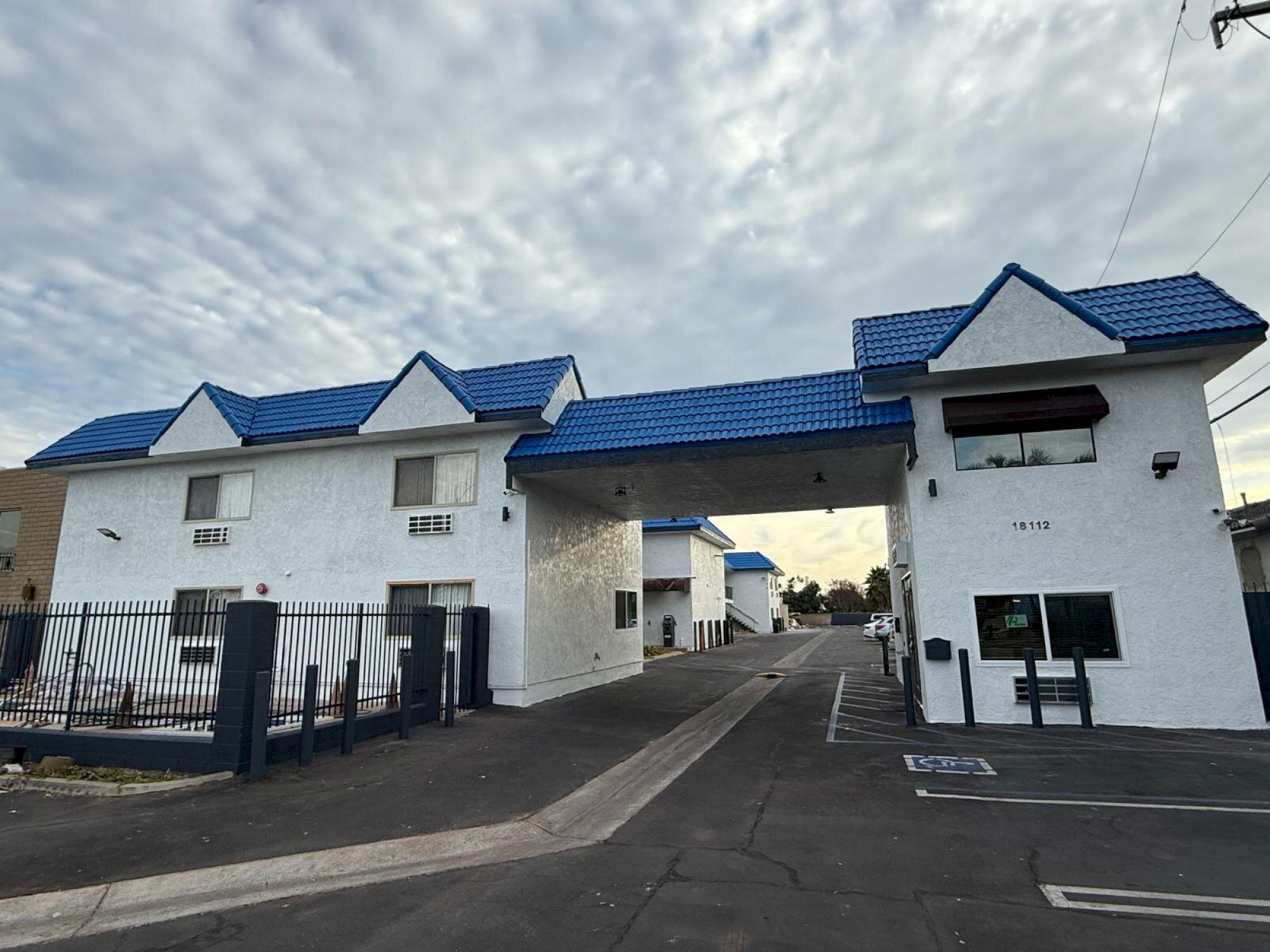 The image shows a white building complex with blue roofs, a gated entry, and parking spaces. The sky is cloudy.