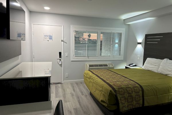 A modern hotel room with a bed, TV, desk, and chair. The floor is wooden, and there is a large window with blinds.