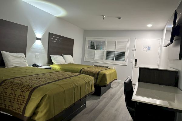 The image shows a modern hotel room with two beds, a desk, a wall-mounted TV, and minimal decor in a bright, clean setting.