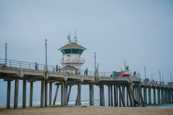 A coastal pier extends over the ocean with a lifeguard tower in view, surrounded by lampposts and some people walking along.