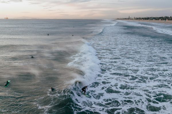 Surfers riding waves near a sandy beach at sunset with a distant view of an oil rig and a line of palm trees along the coast.