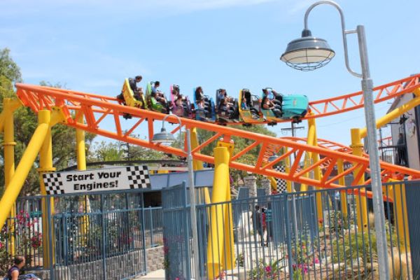 A roller coaster with riders is seen on an orange track above a fenced area. A sign below reads 