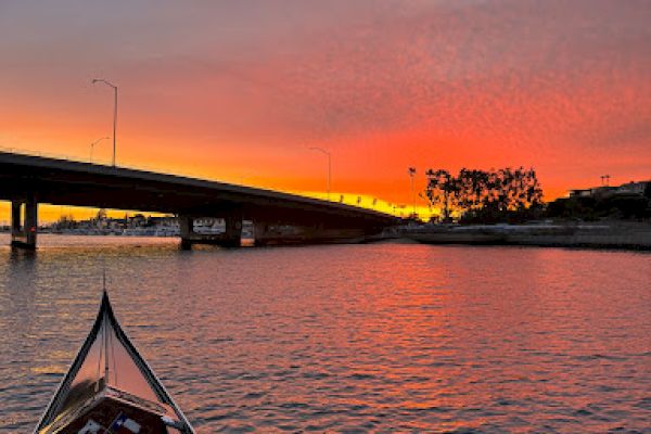 A boat on water approaches a bridge during a vibrant sunset, with colorful skies reflected on the calm surface, creating a serene scene.