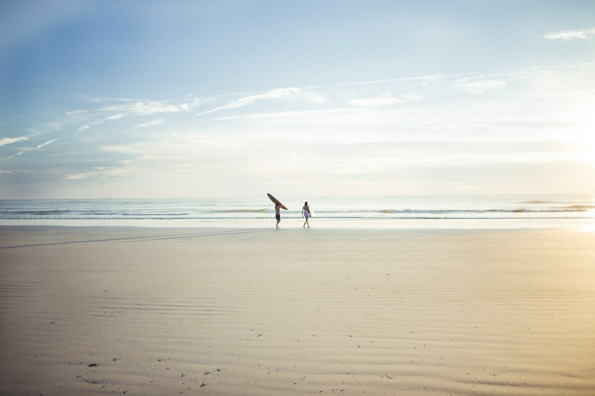 A serene beach scene with two people walking toward the ocean, one carrying a surfboard, under a bright sky.