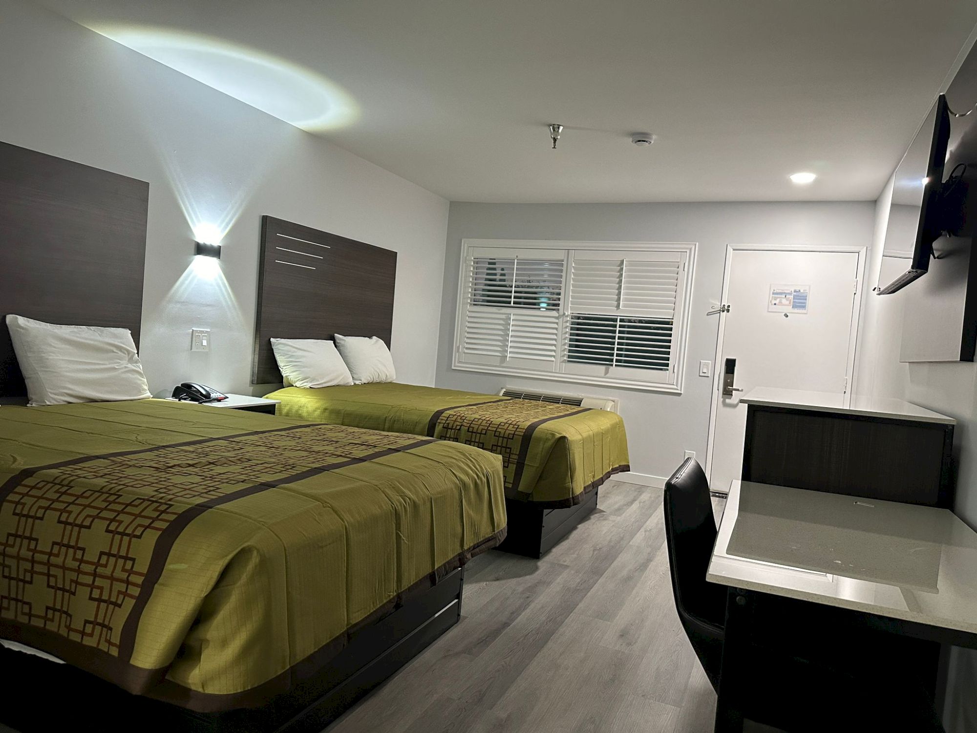 The image shows a clean hotel room with two beds, a TV, desk, and chair, featuring a modern design and muted colors.