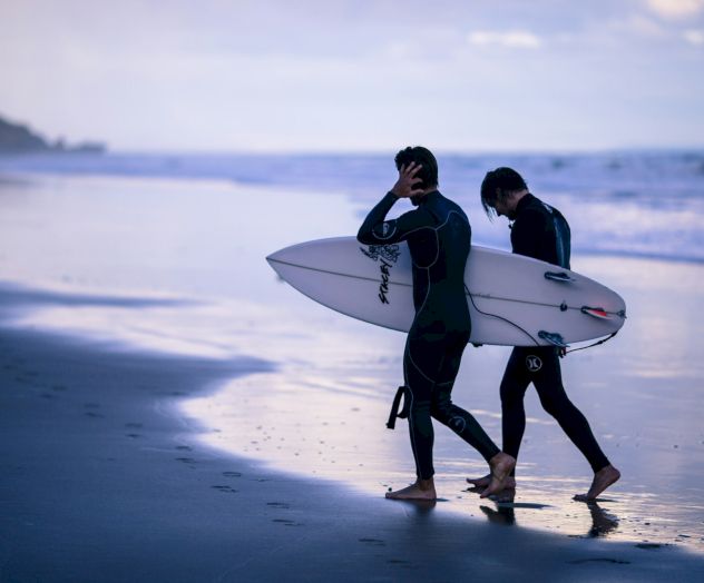 Two people in wetsuits walk along the beach, each carrying a surfboard, with the ocean and a distant headland in the background.