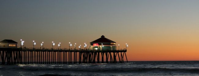 A pier extends into the ocean at sunset, with lights illuminating the structure. The sky transitions from orange to deep blue.