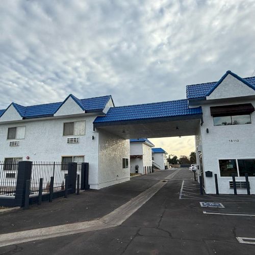 A motel-like building with blue roofs, white walls, a gated area, and a passage leading to a parking lot or inner area.