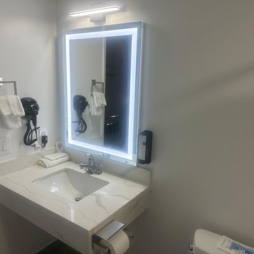 A modern bathroom with a lit mirror above a sink, a hairdryer on the wall, towels, and a toilet paper holder visible.