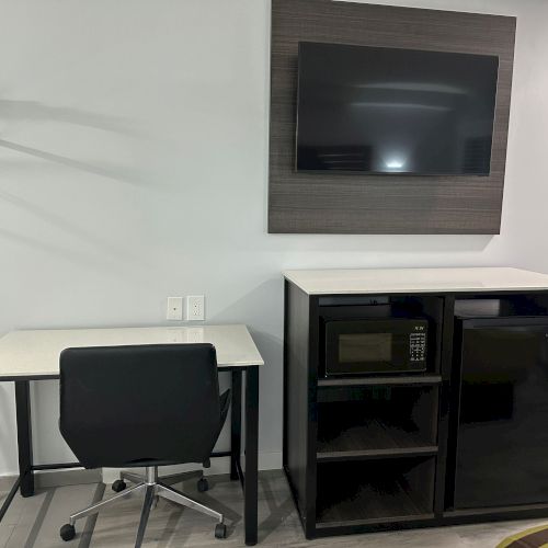 The image shows a desk with a chair, mounted TV, and a cabinet with a microwave and mini fridge in a room.