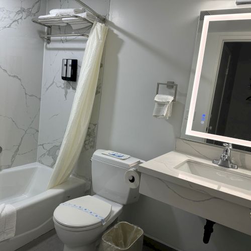 A clean, modern bathroom with a bathtub, toilet, sink, and illuminated mirror. It has a towel rack and marble-patterned walls.