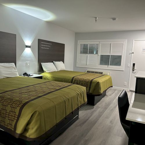 The image shows a hotel room with two beds, a desk, and a chair. The room is minimally decorated and well-lit.