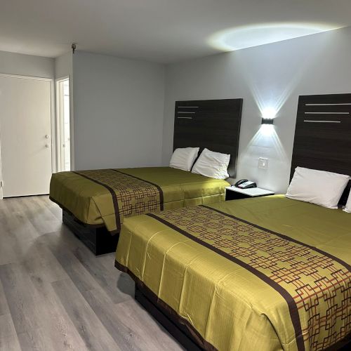 The image shows a hotel room with two double beds, green bedspreads, modern wooden headboards, a nightstand, and light gray walls.