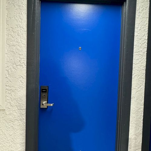 The image shows a vibrant blue door with a modern electronic lock, surrounded by a dark frame against a textured white wall.