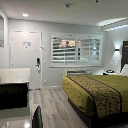 A modern hotel room with a bed, desk, chair, and TV. The room is lit with soft lighting and features a window with blinds.