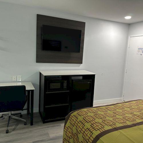 A hotel room with a bed, desk and chair, TV mounted above a cabinet, white walls, and a door.