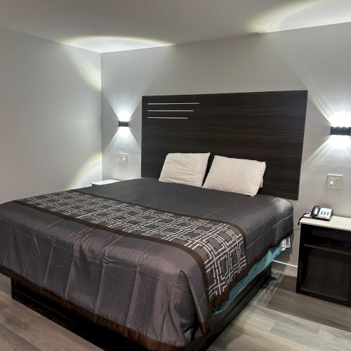 A modern hotel room with a neatly made bed, two pillows, wall-mounted lighting, and a nightstand with a phone.