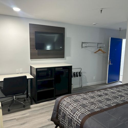 A hotel room with a bed, wall-mounted TV, desk, chair, fridge, and open closet with hangers, and a visible blue door.