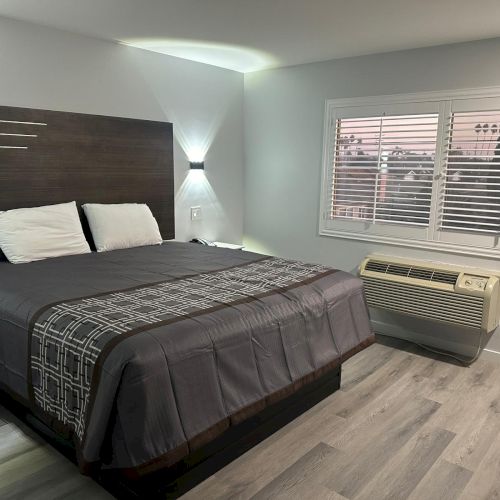 A modern bedroom with a large bed, two pillows, and a stylish headboard. The room features a window with blinds and a wall-mounted AC unit.